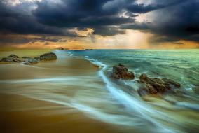 Beautiful landscape of the sandy beach with rocks and white waves, under cloudy sky, at colorful sunset