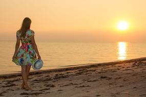 girl in a summer dress with a hat in his hand on the shore of the evening sea