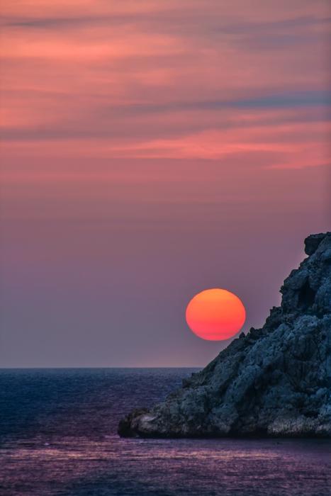 photo of a yellow-pink sun behind the rocks against the background of the evening sky in Greece