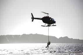 Black and white photo of a person hanging on a rope on a helicopter above the water