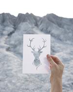 Person holding card with beautiful deer shape, near the grey mountains