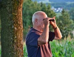 Senior man near the tree, looking in binoculars, among the colorful and beautiful plants