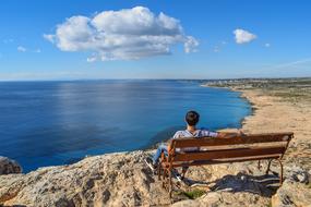 a guy sits on a bench on a rock against the backdrop of the sea