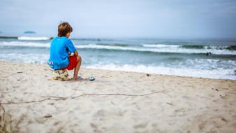 Child sitting on the football ball, on the sandy beach of the ocean with waves