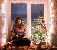 Girl reading book at the window with colorful and beautiful Christmas decorations, Christmas tree and lights, in the at the night