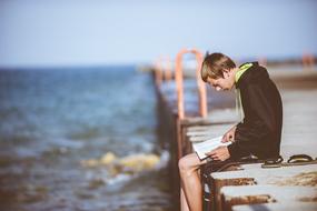 boy reading a book on a pier by the sea in a blurred background