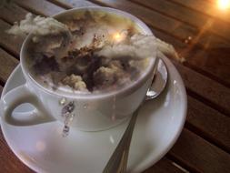 Cup of Cappuccino with clouds and colorful light, on the saucer with spoon, on the wooden table in light