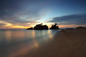 Beautiful landscape of the sandy beach near the rocks with trees, at beautiful and colorful dawn with clouds