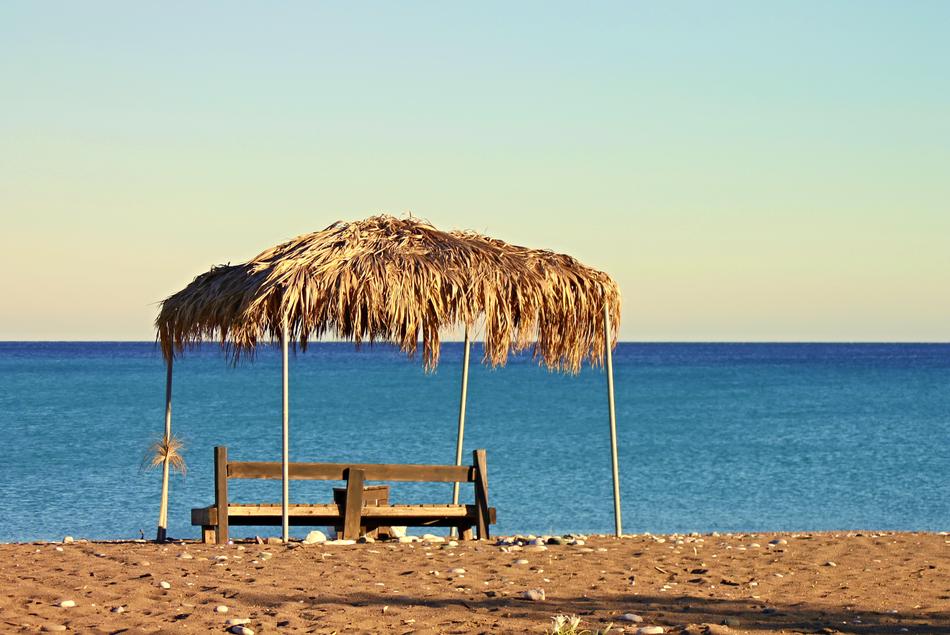 bench covered with construction with the palm leaves, on the sandy beach in sunlight, in Rhodes, Greece, at colorful and beautiful sky on background