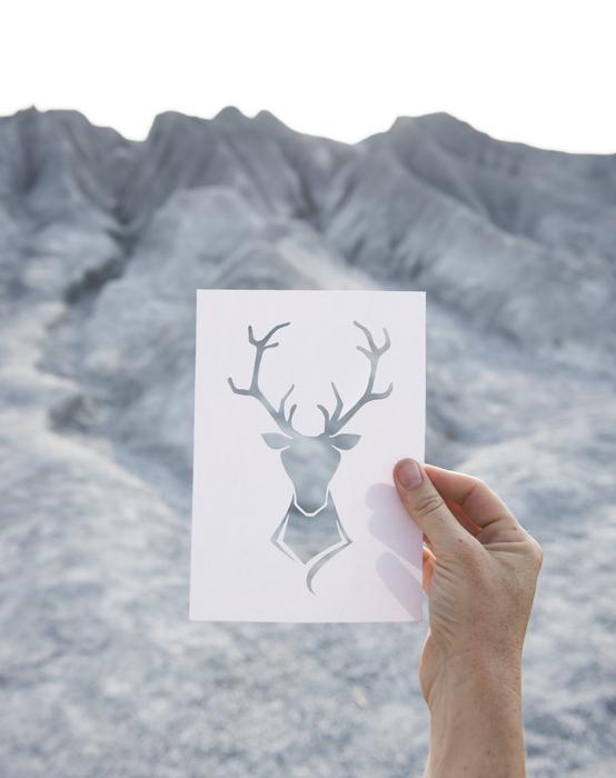 Person holding card with beautiful deer shape, near the grey mountains