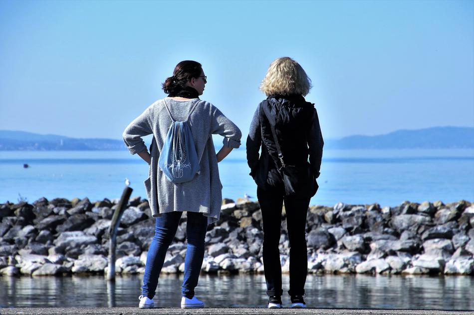 Back view of the girls with bags, standing at the coast of the water, at background with mountains