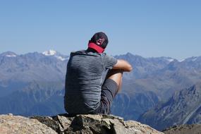 Back view of the guy in cap, sitting on the beautiful mountain among the other mountains with snow