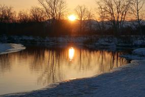 Beautiful landscape with the river, among the snowy shore with plants, at colorful and beautiful sunset