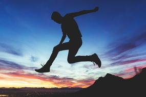 Silhouette of the guy jumping near the silhouettes of mountains in the valley, at colorful and beautiful sunset among the clouds