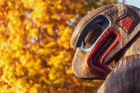 Colorful, wooden totem near the beautiful tree with yellow leaves in sunlight, in the fall