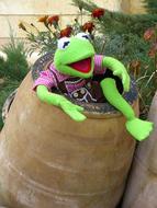 Green Kermit Frog soft toy on a barrel in the garden with red flowers