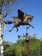 Beautiful, bronze Pegasus, weather gage statue near the green trees, at blue sky on background