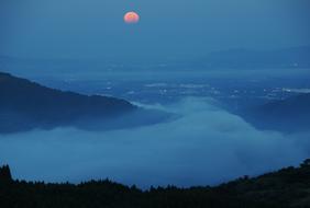 Beautiful night view with the clouds behind the trees, among the mountains, at beautiful and colorful Moon in the sky