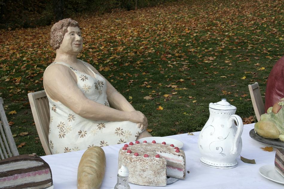 Sculpture if the lady at the served table, near the green grass with colorful leaves