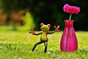 figurine of a funny frog on the grass near a vase with a flower