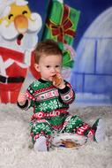 child boy in Christmas clothing eats Cookie on carpet