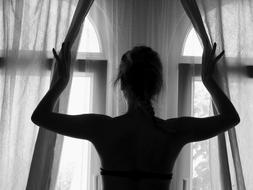 Black and white photo with the silhouette of a woman, opening the curtains of the window