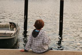 Back view of a child sitting, on the beautiful shore of the sea, with boat