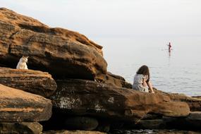 Person, doing stand up paddling in the water, near the beautiful, rocky shore, with the colorful and cute cat and girl