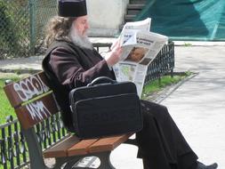 photo of a priest with a newspaper sits on a bench