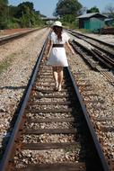 girl in a white dress and hat walks on the railroad tracks