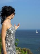 a girl in a gray dress on the coast of Greece points to a sailing ship