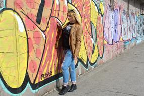 girl in a brown leather jacket stands against a wall with graffiti