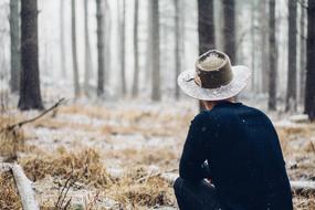 man in wide hat sits on frosted grass in forests