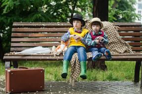 Children in hats, sitting on the bench, among the colorful plants, with the wet pavement, in autumn