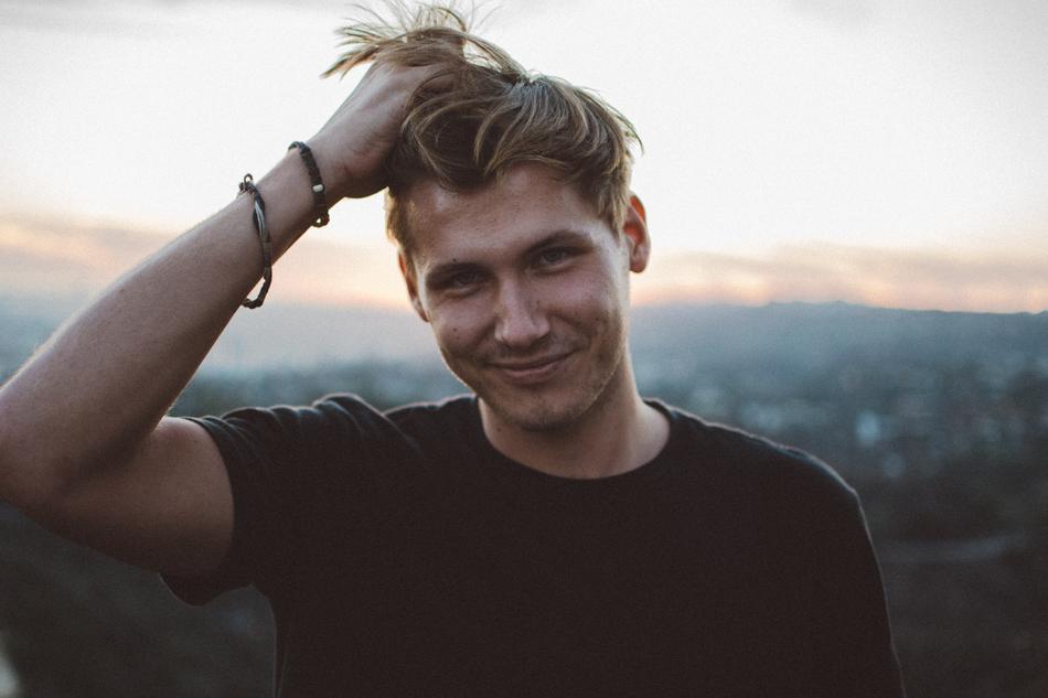 Portrait of the smiling, blonde man in black t-shirt, with the bracelets, at colorful and beautiful background with the mountains