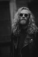 Black and white photo with the portrait of the man in leather jacket, with beard, sunglasses and curly hair