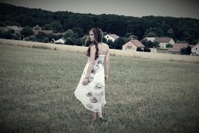 Black and white photo of the beautiful girl in dress, on the beautiful field near the houses, in the village