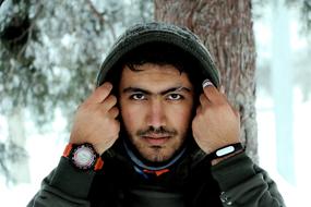 Portrait of the bearded man with wrist watch and bracelet, covered in hood, among the beautiful, snowy forest