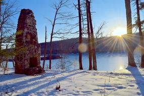 Beautiful, snowy landscape with the stone structure among the trees, near the water in sunlight, in the winter