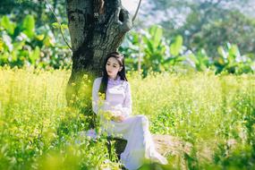 Asian girl, near the tree, among the beautiful and colorful plants on the meadow, in sunlight