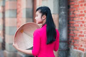 Back view of the smiling girl in pink clothing, with the hat, near the brick wall