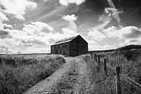 Black and white landscape with the old house near the road with fence, among the plants, under the sky with clouds