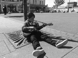 Black and white photo of the sitting boy, playing on the guitar, on the street