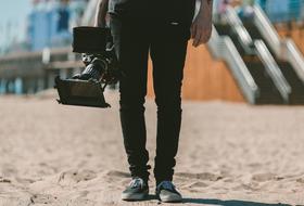 Legs of the person, with the camera, on the sand near the stairs and buildings, in sunlight