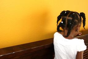 Back view of the cute girl with the braids, in white dress, near the yellow wall