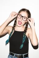 Portrait of the girl, with the colorful braids and glasses, showing tongue, in light, at white background