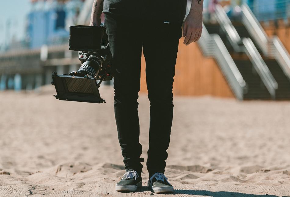 Legs of the person, with the camera, on the sand near the stairs and buildings, in sunlight