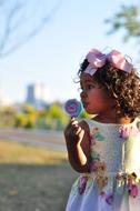 Cute girl with the pink ribbon on the curly hair, in the colorful and beautiful dress, with the colorful lollipop