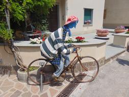 Colorful sculpture of the cyclist, on the retro bike, near the green trees, colorful and beautiful flowers and building