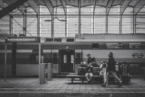 Black and white photo of with the people and train, on the railway station, with the bench